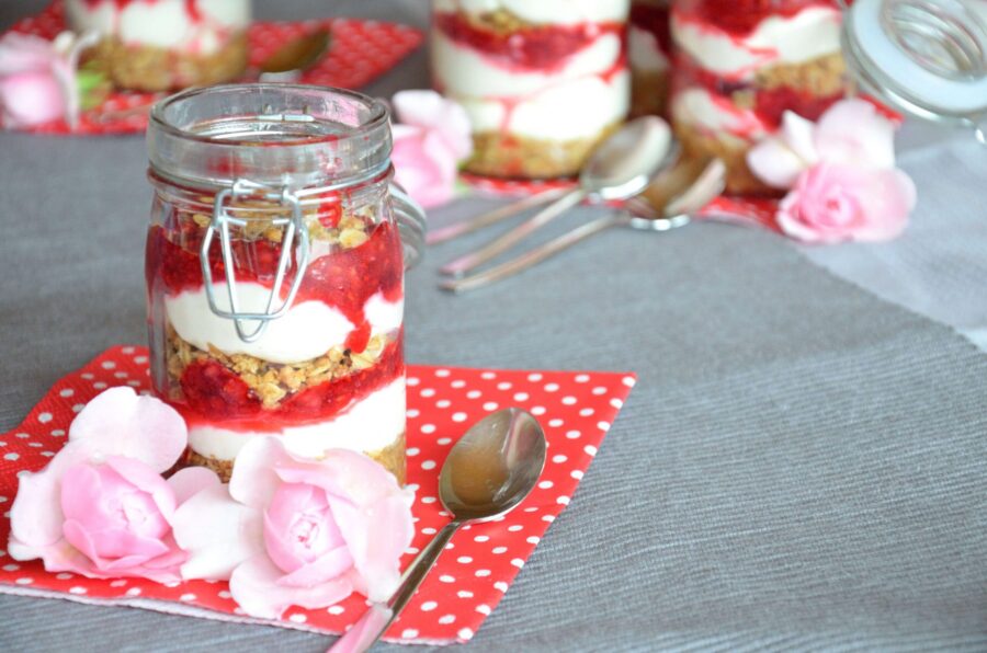 Himbeer-Cheesecake im Glas auf einer roten Serviette mit weißen Punkten, dahinter weitere Gläser mit Himbeer-Cheesecake, von vorne
