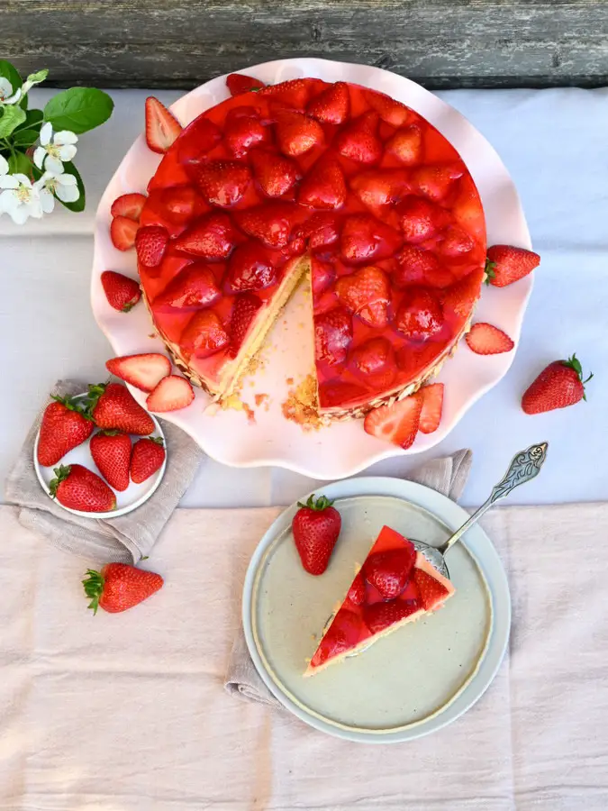 Erdbeertorte mit Vanillecreme, 1 Stück abgeschnitten vorne auf einem Teller, hinten die Torte auf rosa Tortenplatte