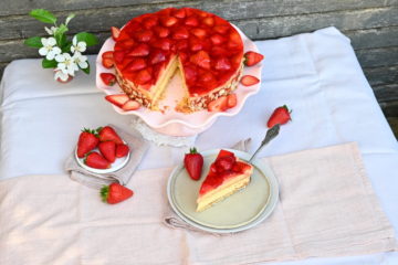 Erdbeertorte mit Vanillecreme, 1 Stück abgeschnitten vorne auf einem Teller, hinten die Torte auf rosa Tortenplatte