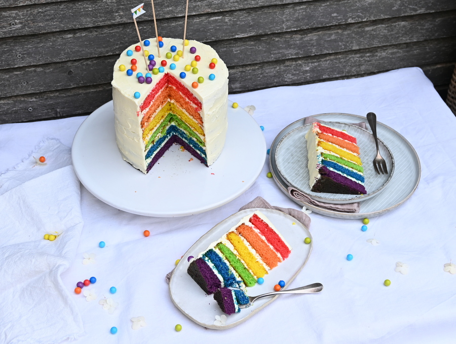 Regenbogen Torte: Vorne 2 Stück Torte auf Tellern, hinten die Torte