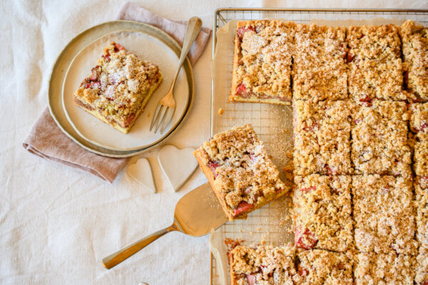 Rhabarberkuchen mit Mandeln auf einem Backgitter, ein Stück wird herausgehoben, von oben
