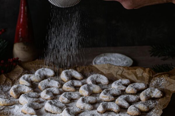 Vanillekipferl auf Backgitter, die geraade mit Staubzucker bestreut werden, Dark & Moody