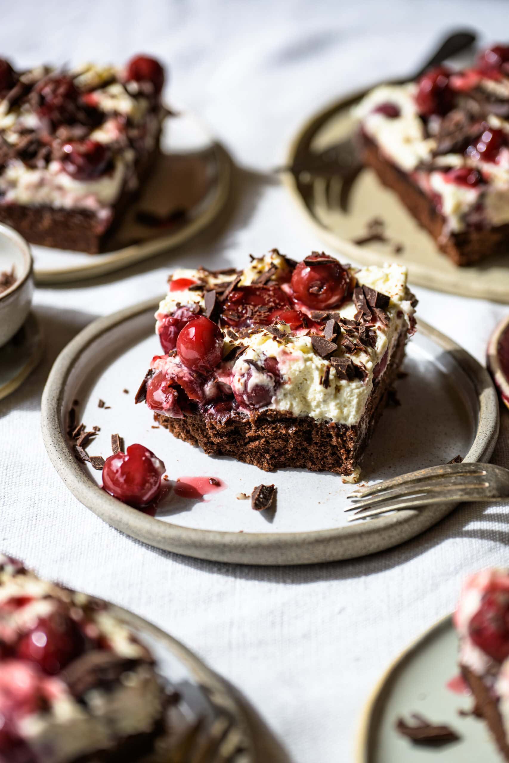 Schwarzwälder Kirschtorte vom Blech, zentral 1 Stück auf einem Teller, runherum sieht man weitere Stück, von schräg vorne