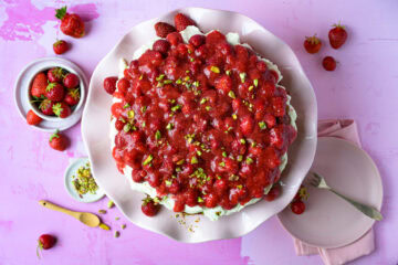 Erdbeertorte mit Frischkäsecreme auf rosa Tortenplatte auf rosa Untergrund, rundherum verteilt Erdbeeren, von oben