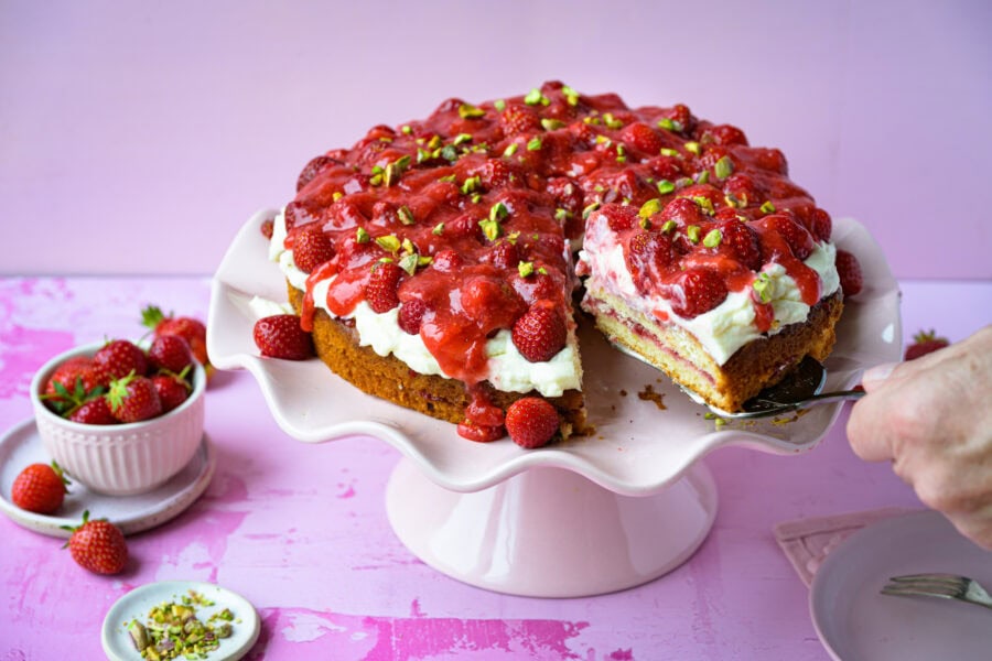 Erdbeertorte mit Frischkäsecreme auf rosa Tortenplatte, angeschnitten, 1 Stück wird gerade herausgehoben