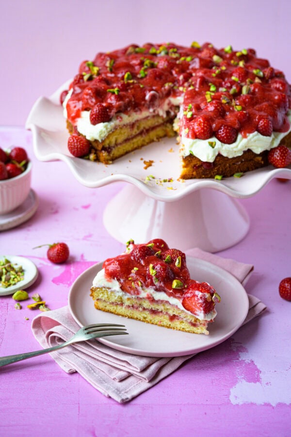 Erdbeertorte mit Frischkäsecreme auf rosa Tortenplatte, angeschnitten, davor ein Stück Torte auf rosa Teller