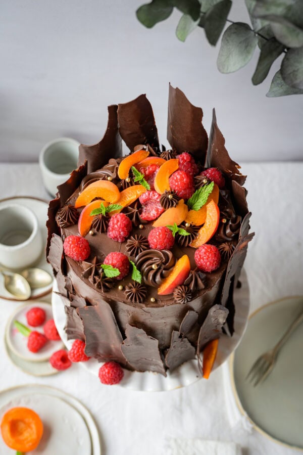 Hohe Schokoladentorte mit einer Deko aus Schokotupfen, Himbeeren und Marillen auf einer weißen Tortenplatte, von oben