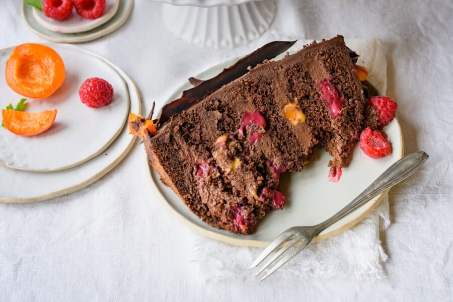 Hohe Schokoladentorte mit einer Deko aus Schokotupfen, Himbeeren und Marillen, ein Stück auf einem Teller