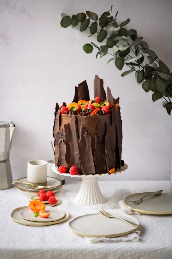 Hohe Schokoladentorte mit Himbeeren und Marillen auf einer weißen Tortenplatte, von vorne