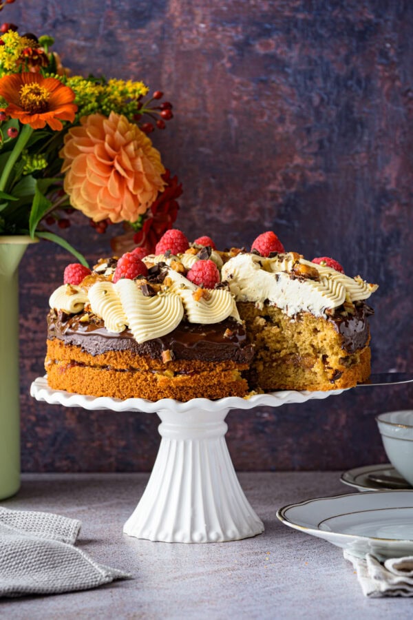 Kaffeebuttercremetorte mit Pekan-Salzkaramell auf einer weißen Tortenplatte, Tortenheber hebt ein Stück heraus, hinten ein bunter Blumenstrauß, von schräg vorne,