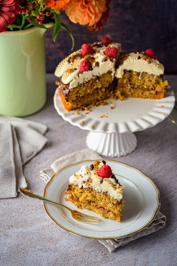 Kaffeebuttercremetorte mit Pekan-Salzkaramell auf einer weißen Tortenplatte, vorne 1 Stück auf einem Teller, im Hintergrund ein bunter Blumenstrauß