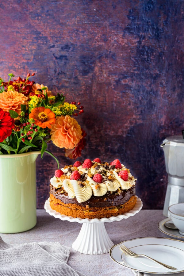 Kaffeebuttercremetorte mit Pekan-Salzkaramell auf einer weißen Tortenplatte, von schräg vorne, hinten ein bunter Blumenstrauß