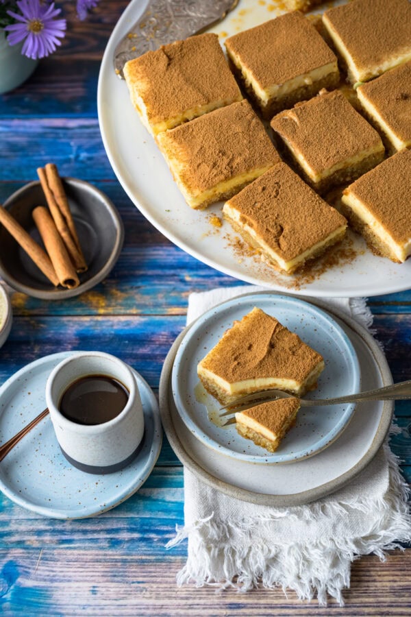 Politiko Dessert: Griechischer Grießkuchen mit Zimt: ein Stück auf einem Teller, mit Gabel drin, von schräg vorne, hinten weitere Stück auf einer Tortenplatte